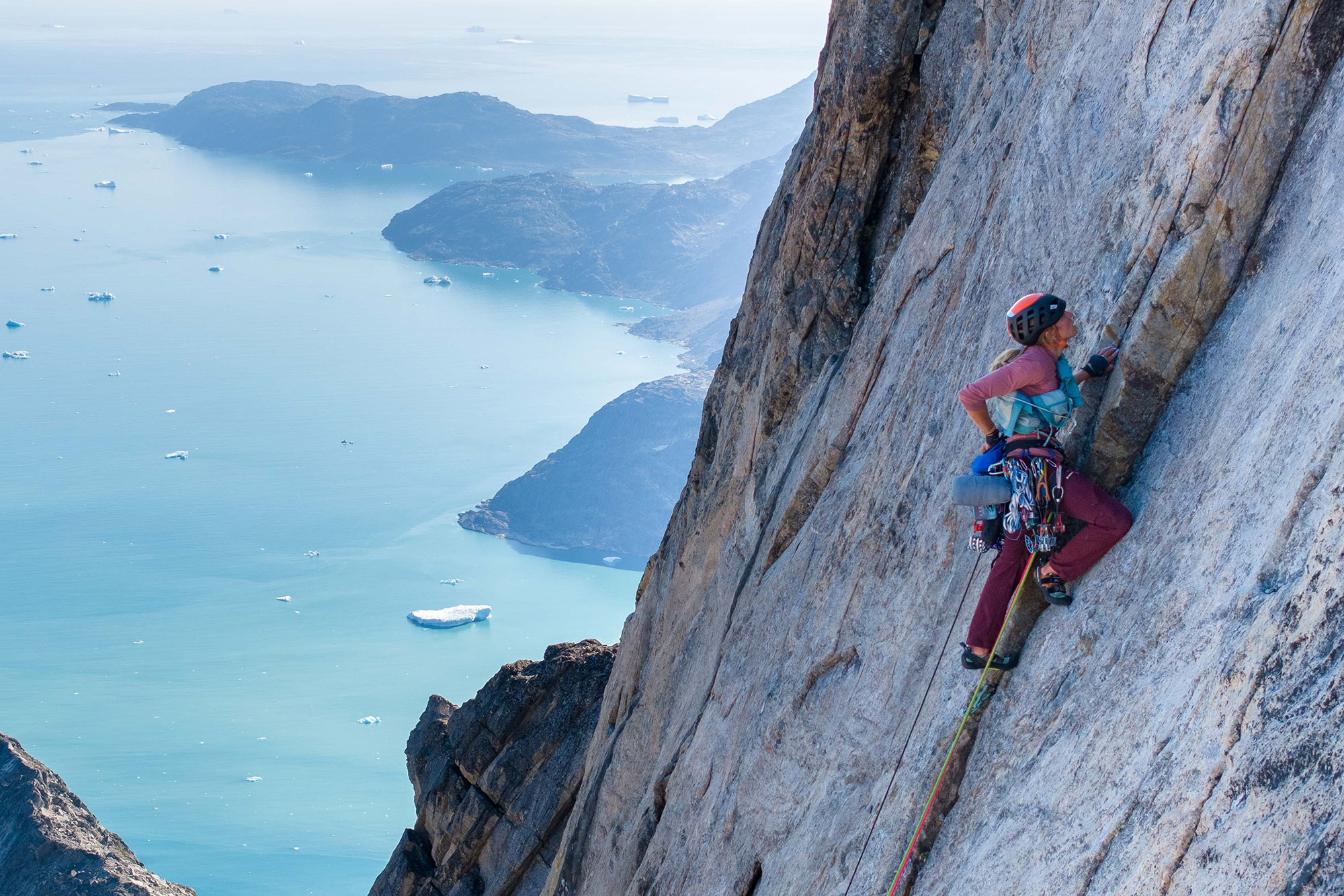 Júlia aparece escalando una preciosa pared de roca muy escarpada, con pantalón morado y camiseta azul de tirantes, casco rojo y arnés lleno de material de escalada. De fondo y bajo ella una impresionante imagen de fiordo groenlandés con cascotes de hielo flotando sobre el agua