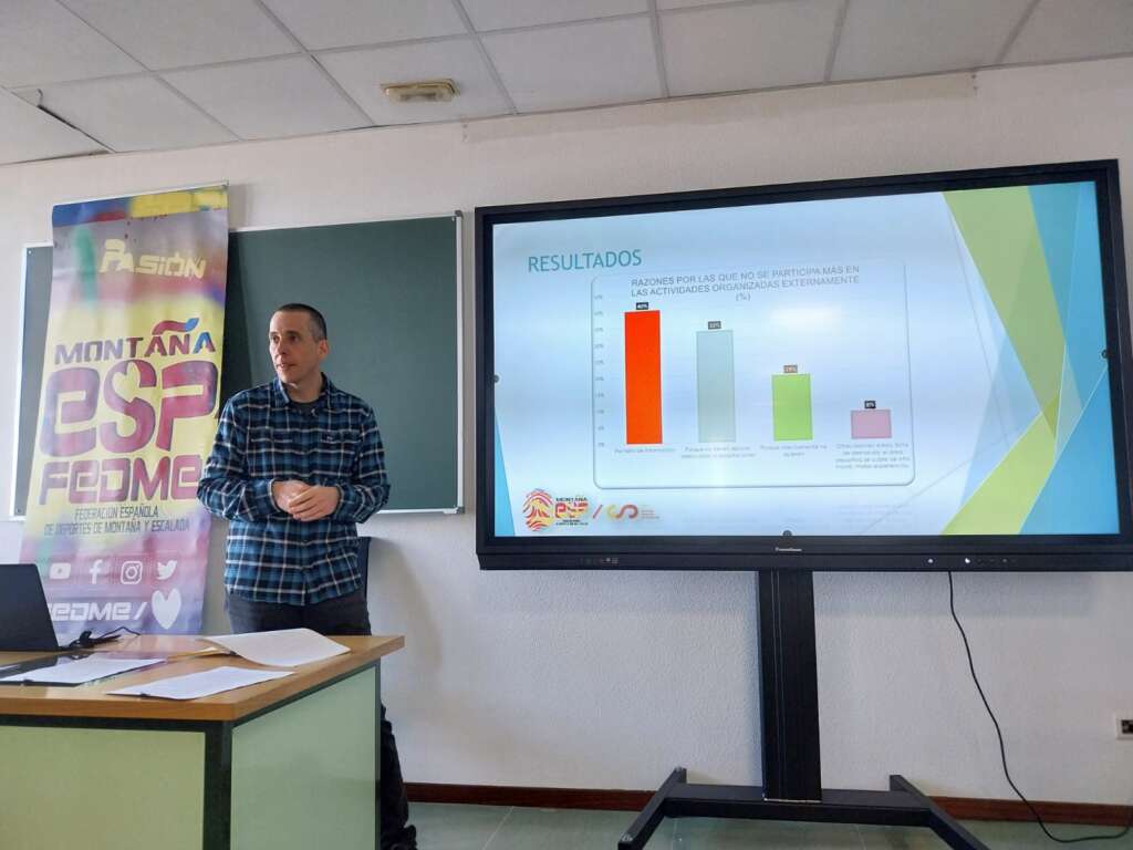 Foto 1: La fotografía muestra a un hombre en el interior de un aula de la Facultad de Ciencias de la Actividad Física y del Deporte. El hombre está de pie exponiendo una explicación junto a una pizarra que muestra un gráfico de barras horizontales.